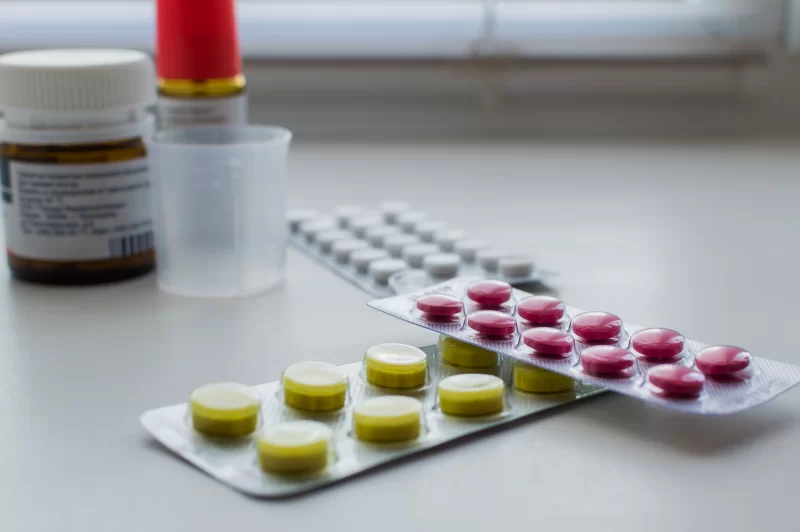 Лекарства на подоконнике: блистеры с таблетками, бутылочка и мерный стаканчик