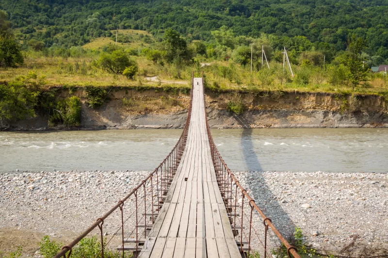 Живописный висячий мост над бурной горной рекой, ведущий к лесу