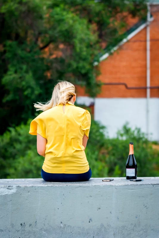 Девушка в желтом сидит на стене с бутылкой вина и телефоном, задумчивый момент