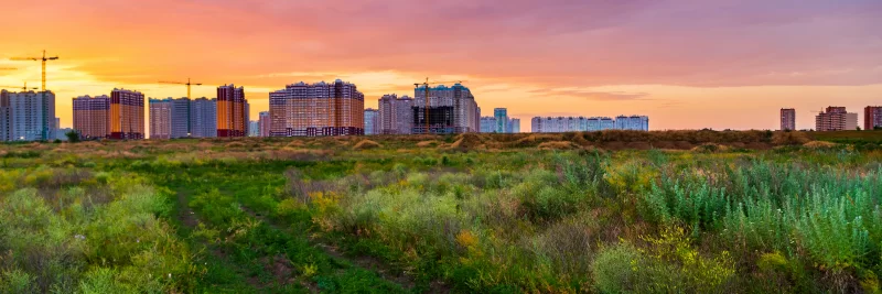 Закат над строящимся городом с зелёной растительностью на переднем плане
