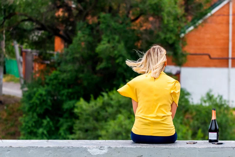 Одинокая девушка в желтой блузке сидит на стене с бутылкой вина и телефоном