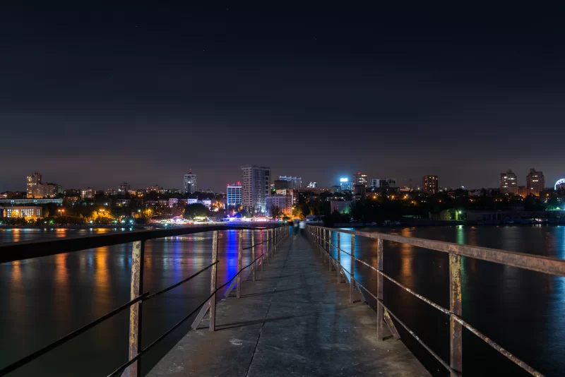 Старый длинный металлический пирс над рекой. Ночной городской пейзаж.